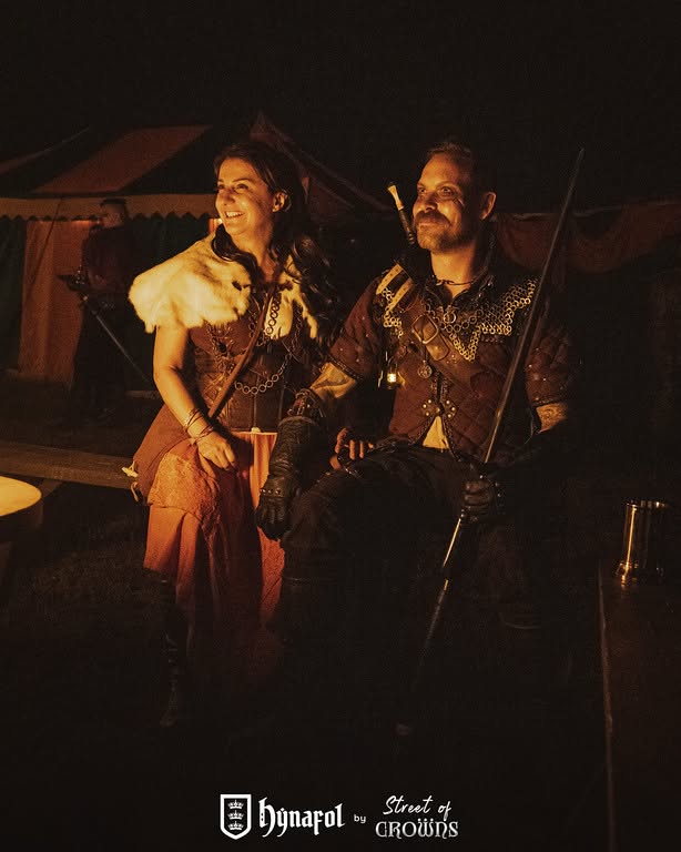 Talia and Korsin together at Hynafol, their faces and costumes lit by the warm glow of the fire.